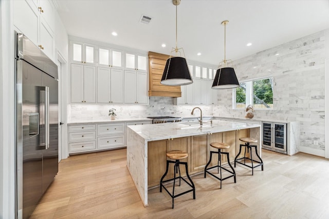 kitchen featuring a kitchen island with sink, built in fridge, white cabinets, sink, and light stone counters