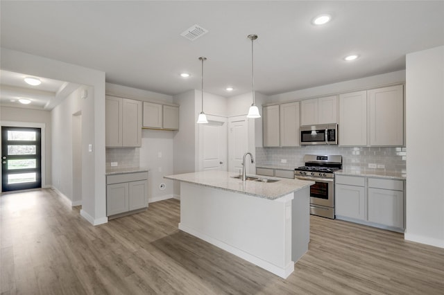 kitchen with appliances with stainless steel finishes, light wood-type flooring, light stone counters, a kitchen island with sink, and sink