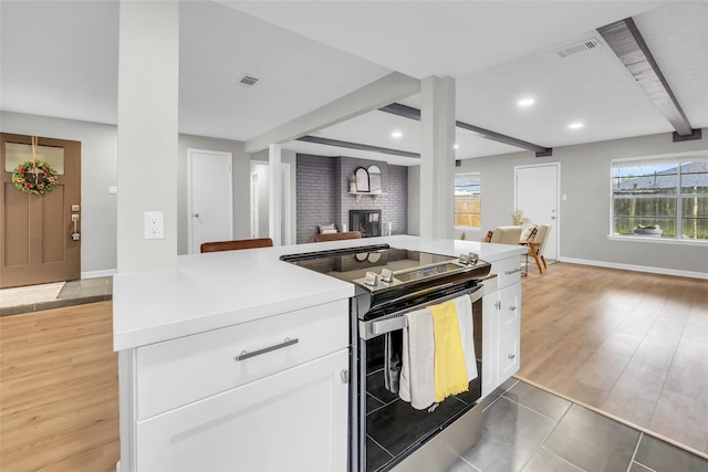 kitchen with beam ceiling, black range with electric stovetop, a brick fireplace, white cabinets, and light wood-type flooring