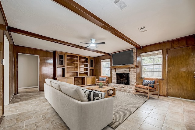 living room with a fireplace, a textured ceiling, ceiling fan, wooden walls, and beamed ceiling