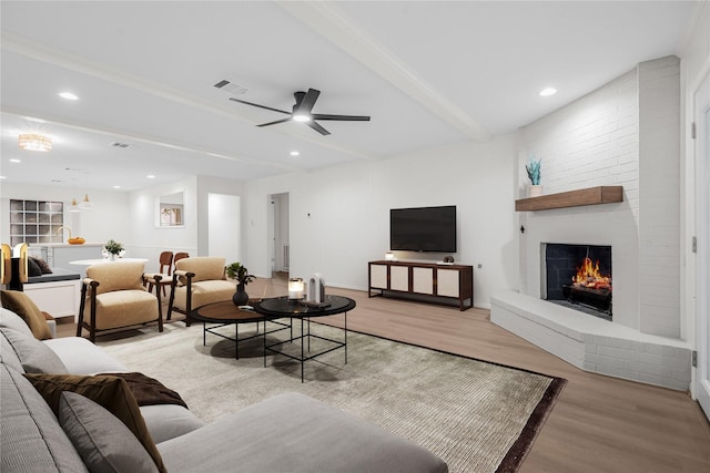 living room featuring ceiling fan, beamed ceiling, light hardwood / wood-style floors, and a brick fireplace