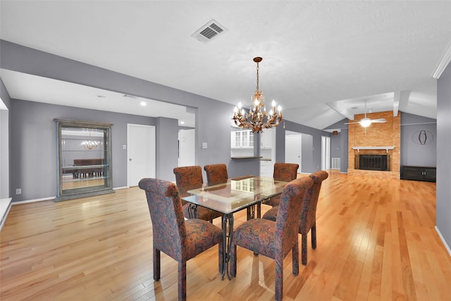 dining space with a brick fireplace, ceiling fan with notable chandelier, light wood-type flooring, and vaulted ceiling