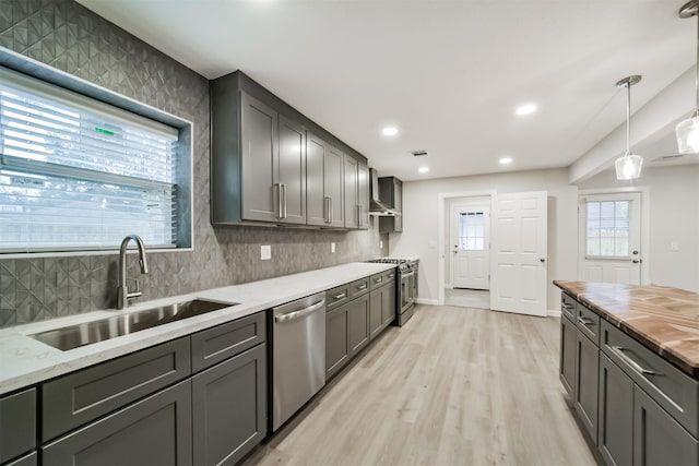 kitchen with pendant lighting, sink, light hardwood / wood-style flooring, wall chimney exhaust hood, and stainless steel appliances