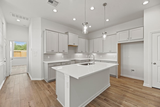 kitchen with light wood-type flooring, tasteful backsplash, sink, decorative light fixtures, and a center island with sink