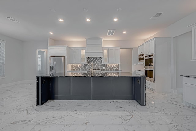 kitchen featuring decorative backsplash, a kitchen island with sink, stainless steel appliances, white cabinets, and dark stone counters