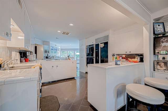 kitchen with tile countertops, kitchen peninsula, white cabinets, tile patterned flooring, and backsplash