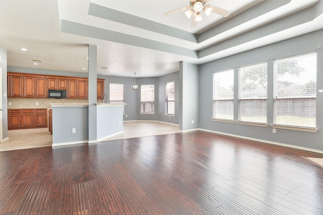 unfurnished living room with a tray ceiling, sink, ceiling fan with notable chandelier, and light wood-type flooring