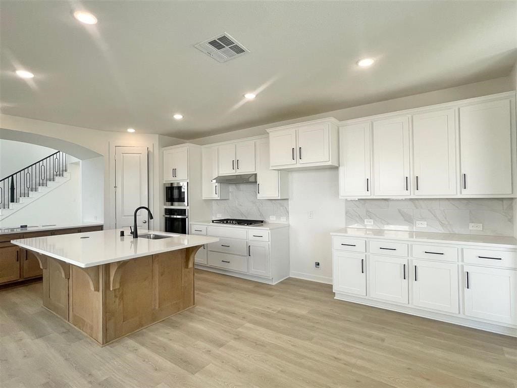 kitchen with decorative backsplash, light wood-type flooring, stainless steel appliances, white cabinets, and an island with sink