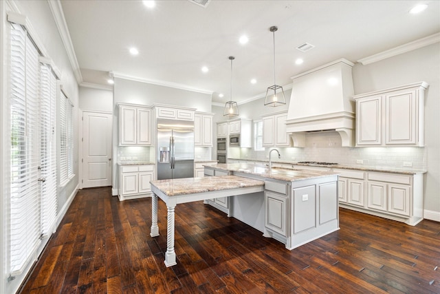 kitchen with built in appliances, premium range hood, hanging light fixtures, an island with sink, and tasteful backsplash