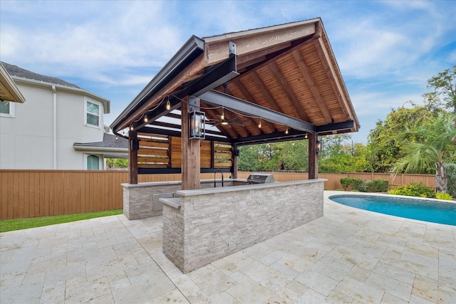 view of patio featuring an outdoor kitchen, a gazebo, a bar, a fenced in pool, and ceiling fan