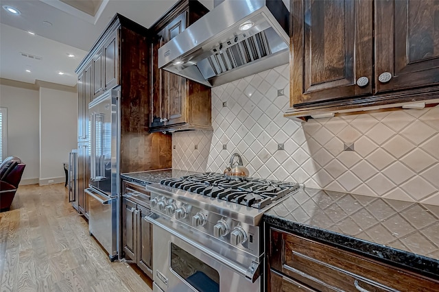 kitchen with light wood-type flooring, backsplash, premium appliances, wall chimney range hood, and dark stone countertops