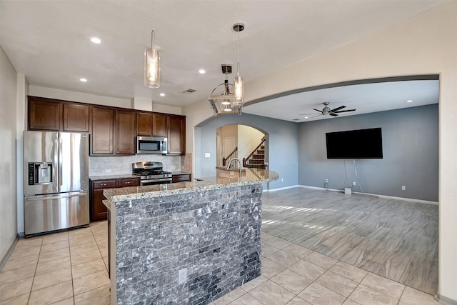 kitchen with dark brown cabinetry, light stone countertops, hanging light fixtures, stainless steel appliances, and a kitchen island with sink