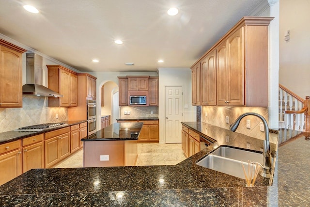 kitchen with wall chimney range hood, sink, dark stone countertops, ornamental molding, and appliances with stainless steel finishes