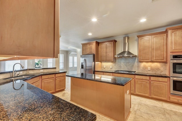 kitchen with dark stone counters, wall chimney range hood, sink, appliances with stainless steel finishes, and a kitchen island