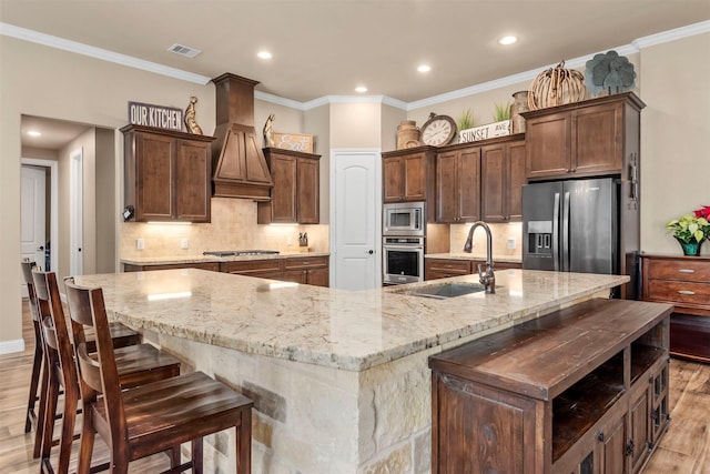 kitchen featuring light wood finished floors, custom range hood, appliances with stainless steel finishes, a large island, and a sink