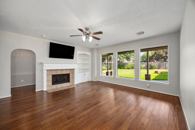 unfurnished living room with ceiling fan, built in shelves, dark wood-type flooring, and a tiled fireplace