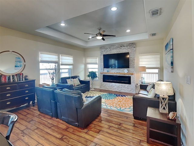 living room with wood-type flooring, a raised ceiling, ceiling fan, and a stone fireplace