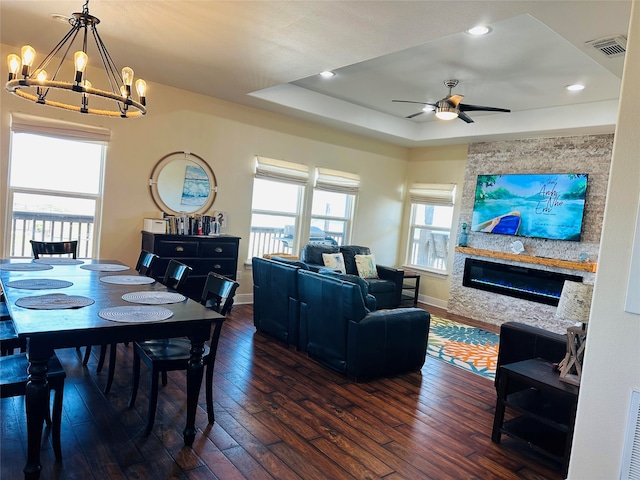 living room with a raised ceiling, ceiling fan with notable chandelier, dark hardwood / wood-style floors, and a stone fireplace