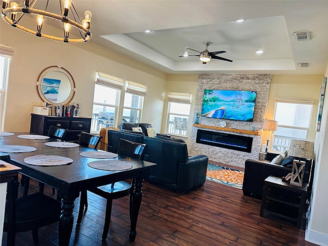 dining space with ceiling fan with notable chandelier, a stone fireplace, dark wood-type flooring, and a tray ceiling