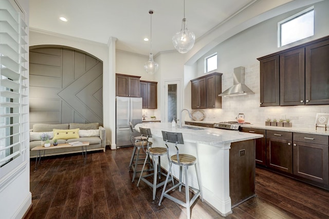 kitchen with dark hardwood / wood-style flooring, wall chimney exhaust hood, a center island with sink, and appliances with stainless steel finishes