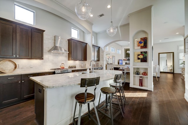 kitchen featuring wall chimney exhaust hood, dark wood-type flooring, sink, decorative light fixtures, and a center island with sink