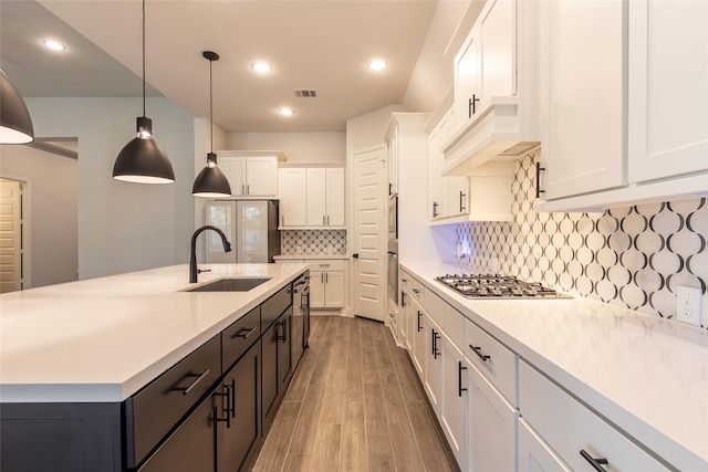 kitchen with appliances with stainless steel finishes, a kitchen island with sink, sink, white cabinetry, and hanging light fixtures