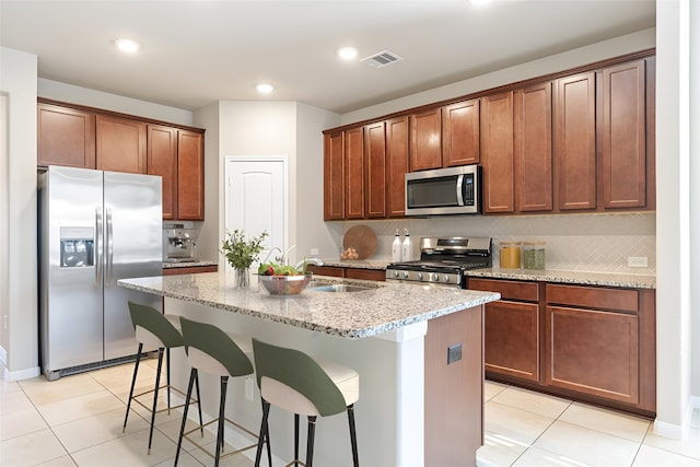 kitchen featuring light stone countertops, appliances with stainless steel finishes, backsplash, a kitchen island with sink, and light tile patterned floors