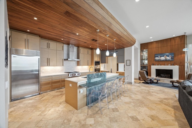 kitchen with pendant lighting, a center island with sink, built in appliances, wall chimney exhaust hood, and tasteful backsplash