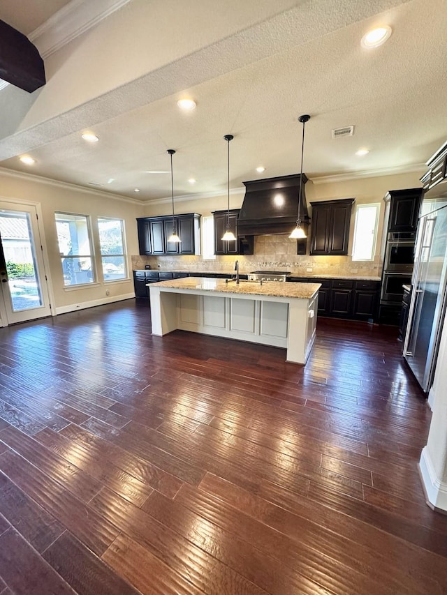 kitchen featuring a center island with sink, backsplash, premium range hood, and hanging light fixtures