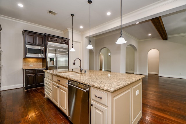 kitchen featuring built in appliances, a kitchen island with sink, dark brown cabinetry, a sink, and decorative light fixtures