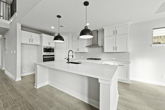 kitchen with light stone counters, stainless steel appliances, sink, wall chimney range hood, and a center island with sink