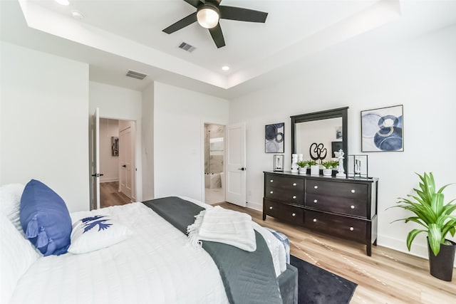 bedroom with ceiling fan, light wood-type flooring, a raised ceiling, and connected bathroom