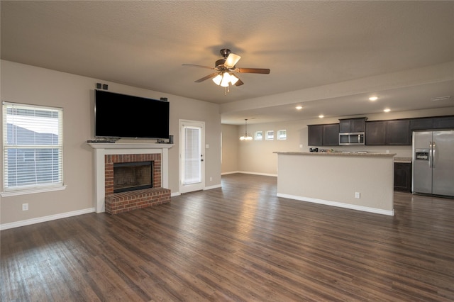 unfurnished living room featuring ceiling fan with notable chandelier, a textured ceiling, dark hardwood / wood-style flooring, and a brick fireplace