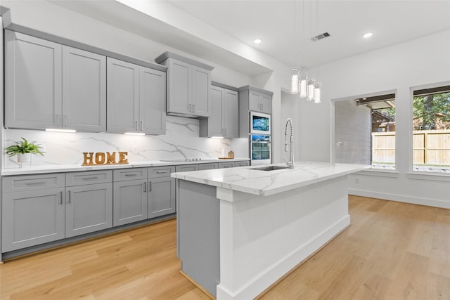 kitchen with a kitchen island with sink, hanging light fixtures, sink, light hardwood / wood-style flooring, and light stone countertops