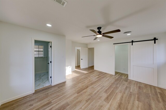 unfurnished bedroom with connected bathroom, a barn door, ceiling fan, and light wood-type flooring