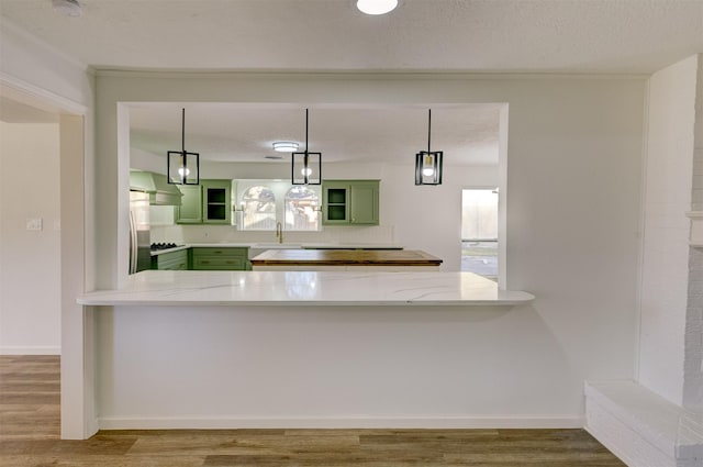 kitchen featuring wall chimney range hood, green cabinets, kitchen peninsula, pendant lighting, and light hardwood / wood-style floors