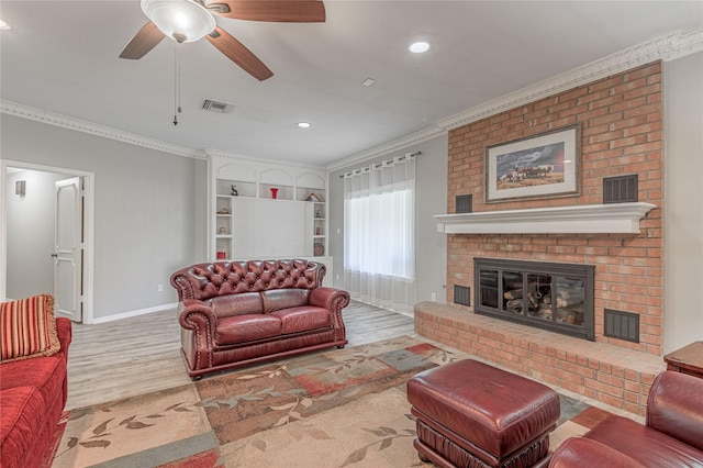 living room with built in shelves, ceiling fan, a brick fireplace, light hardwood / wood-style flooring, and crown molding