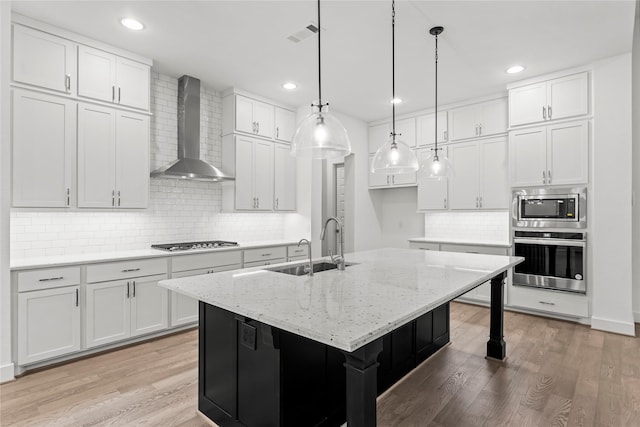 kitchen featuring appliances with stainless steel finishes, wall chimney exhaust hood, a center island with sink, light hardwood / wood-style flooring, and white cabinets