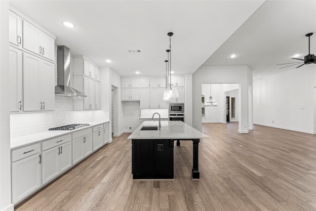 kitchen with white cabinets, wall chimney exhaust hood, light hardwood / wood-style floors, and an island with sink