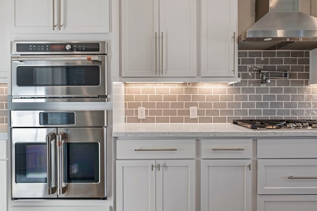 kitchen featuring decorative backsplash, white cabinets, stainless steel appliances, and wall chimney range hood