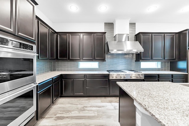 kitchen featuring decorative backsplash, wall chimney range hood, dark brown cabinetry, and stainless steel appliances