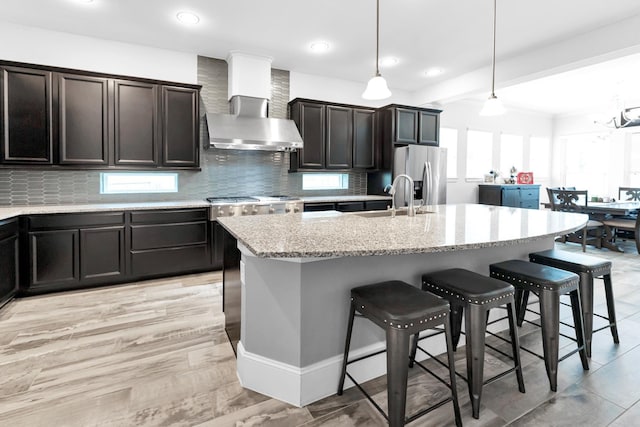 kitchen featuring wall chimney exhaust hood, sink, hanging light fixtures, stainless steel fridge, and a center island with sink