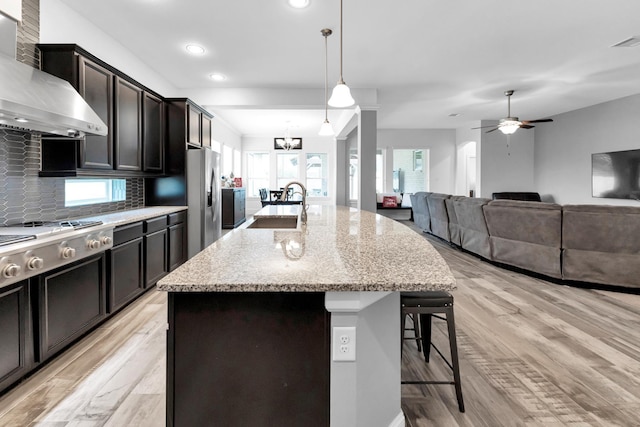 kitchen featuring wall chimney exhaust hood, decorative light fixtures, tasteful backsplash, sink, and a kitchen island with sink