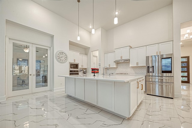 kitchen featuring white cabinetry, a towering ceiling, decorative light fixtures, a center island with sink, and appliances with stainless steel finishes