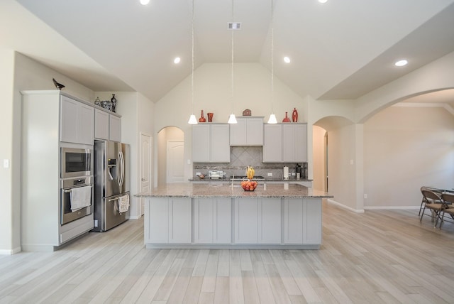 kitchen with stainless steel appliances, light stone counters, light hardwood / wood-style flooring, pendant lighting, and a kitchen island with sink