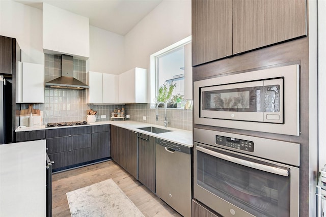 kitchen with sink, wall chimney exhaust hood, appliances with stainless steel finishes, white cabinets, and light wood-type flooring