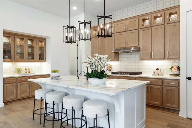 kitchen with a kitchen island with sink, an inviting chandelier, light hardwood / wood-style floors, light stone counters, and stainless steel gas cooktop