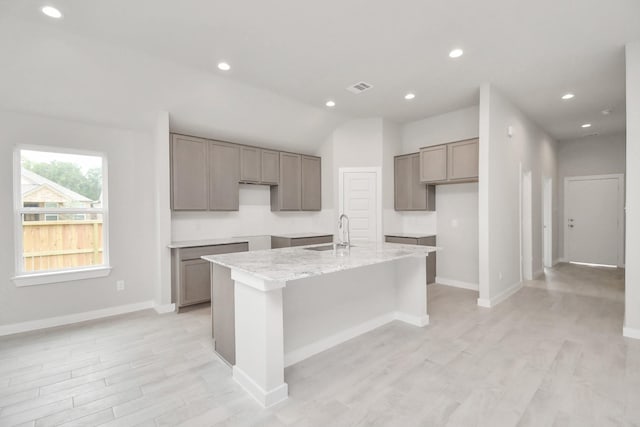 kitchen with light stone countertops, sink, an island with sink, light hardwood / wood-style floors, and vaulted ceiling