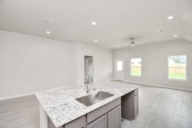 kitchen featuring light wood-type flooring, light stone counters, ceiling fan, sink, and an island with sink