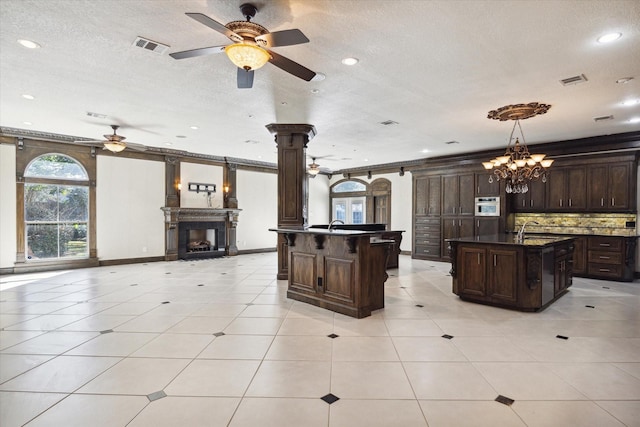 kitchen with dark brown cabinets, ornamental molding, hanging light fixtures, and a kitchen island with sink